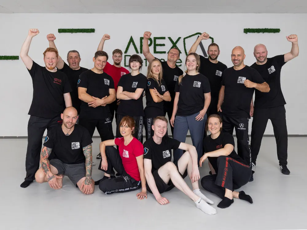 Das Team von Apex Defence posiert in schwarzen Shirts vor der Wand mit dem Apex-Logo, einige recken die Faust nach oben
