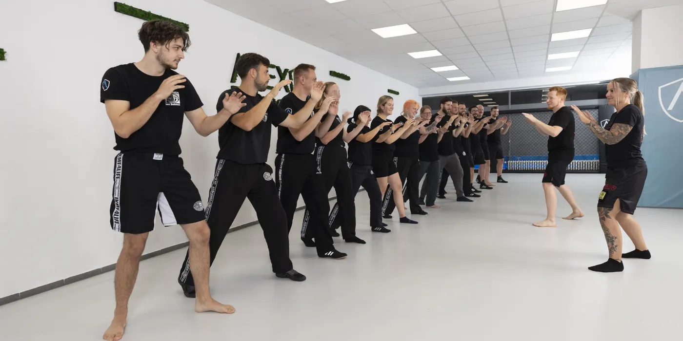 Trainingsgruppe in schwarzer Apex-Defence-Kleidung steht in zwei Reihen in der Kampfhaltung in der hellen Trainingshalle, zwei Trainer stehen ihnen gegenüber